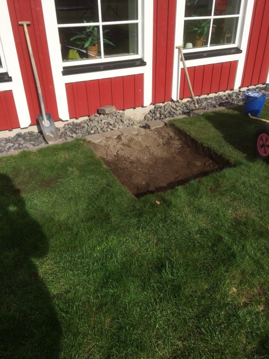 Grävd kvadratisk grop i gräsmatta framför en röd stuga, med en spade lutad mot husväggen.