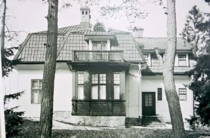 Svartvitt foto av en tvåvåningsvilla med veranda och mansardtak, omgiven av träd, från 1920-talet.