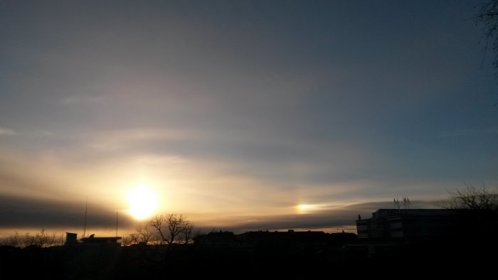 Solnedgång med ljusfenomen, känd som halo, vid sidan av solen, över siluetten av stadens byggnader.