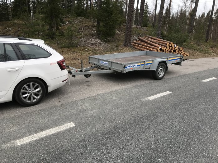 Vit bil kopplad till ett tomt, tvåaxlat släpvagn parkerad på en väg bredvid skog.