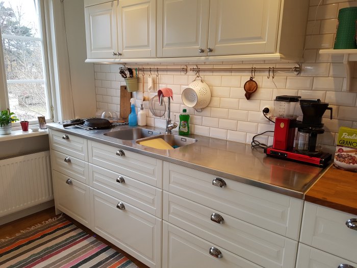 Ett kök med vita skåp och rostfritt stål bänkskiva, kakel, kaffeapparat och inredningsdetaljer.