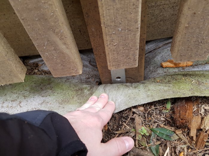 Hand pekar på en insjunken markskruv under vedbod, med synlig glipa där stolpe möter markduken.