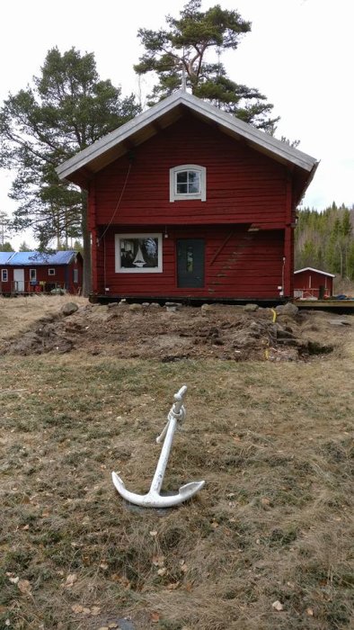 Röd stuga med avlägsnad altan och synlig sandmark, förberedd för ny altanbyggnation, med ankare i förgrunden.