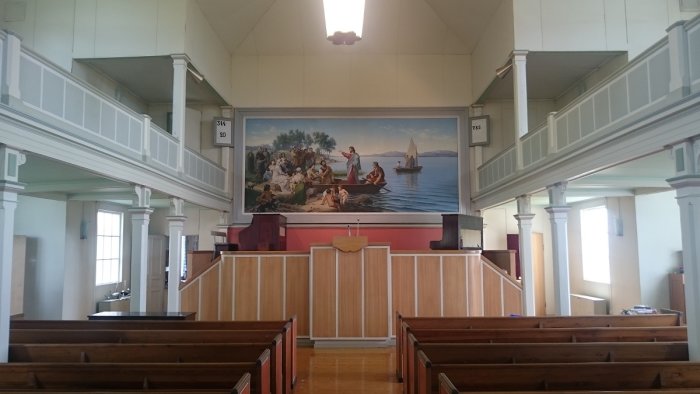 Interiör av en kyrka med bänkar och ett podium framför en stor biblisk målning där Jesus pekar mot åskådaren.