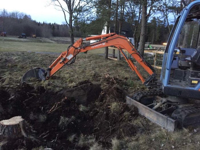 En svetsad ripper på en mindre orangefärgad grävmaskin i skogsmark.