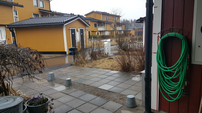 Utsikt över en trädgård med våta stenplattor, krukor och justerbara stolpskor där ett plank ska byggas.