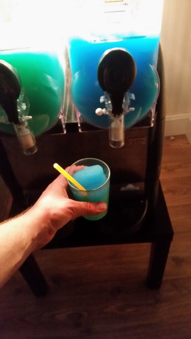 En person håller ett glas med blå slush framför en slushmaskin med två behållare, en grön och en blå, under pausen i kaklingsarbetet.