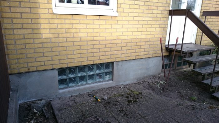 Husvägg med tegelfasad och grund, befintligt altandäck och trappa, mätverktyg på marken.