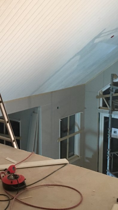 Renoverat rum med nylagt takpanel, gipsväggar, och fönsterinfattningar; byggmaterial och verktyg i förgrunden.