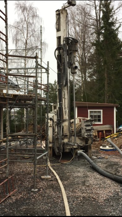 Borrigg i arbete för bergvärmeinstallering på en byggarbetsplats med ställningar och en röd stuga i bakgrunden.