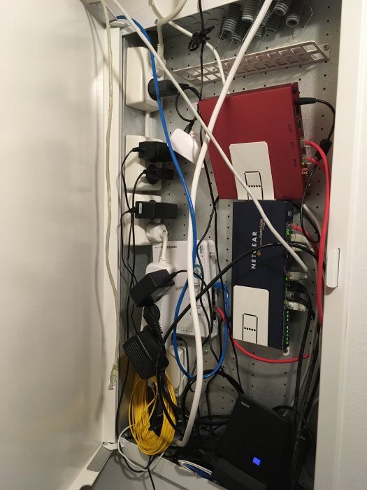 Översikt av rörig nätverksskåp med diverse utrustningar och kablar inklusive en PfSense-låda och en Netgear-switch.