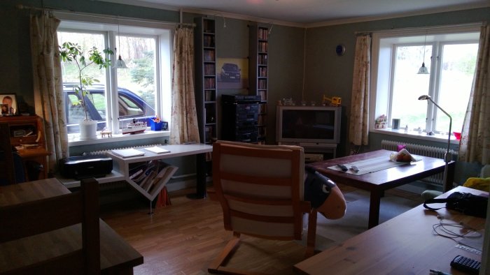 Ett vardagsrum med skrivbord, TV, bokhyllor och stolar, genom fönstret syns en bilparkering.