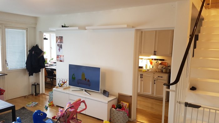 Inredning av ett vardagsrum med vit vägg, kabel-TV, leksaker och utsikt till ett kök bakom samt trappa till höger.