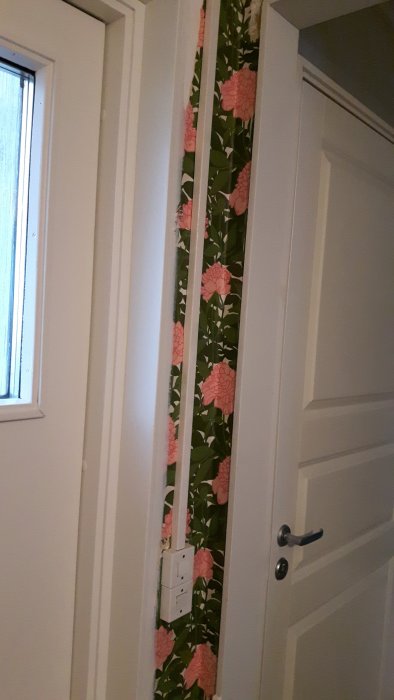 Vägg med synlig kabel och eluttag på tapet med blommönster, bredvid en dörr med handtag.