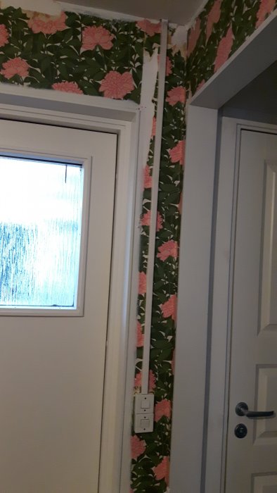 En vägg med synliga elledningar och blommigt tapetmönster, intill ett fönster och en dörr.