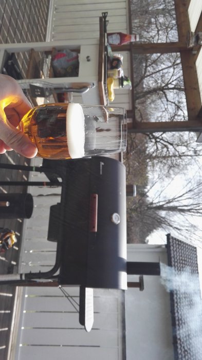 Person som håller en öl framför en rökande grill på en veranda, avslappnad miljö.