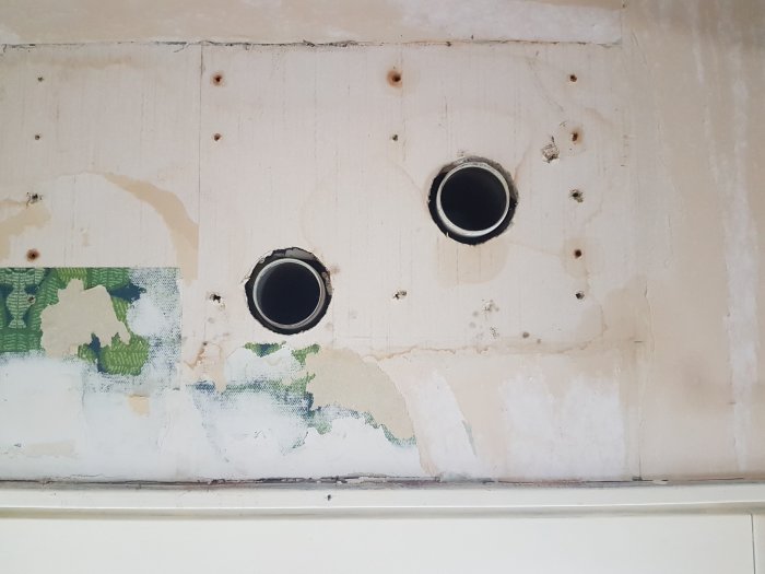 Två genomgående hål i en vit vägg med rester av gammal tapet och spår efter borttagen installation.