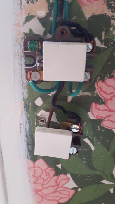 En öppen strömbrytare med lösa och felaktigt sammanlindade kablar på en vägg med blommig tapet.
