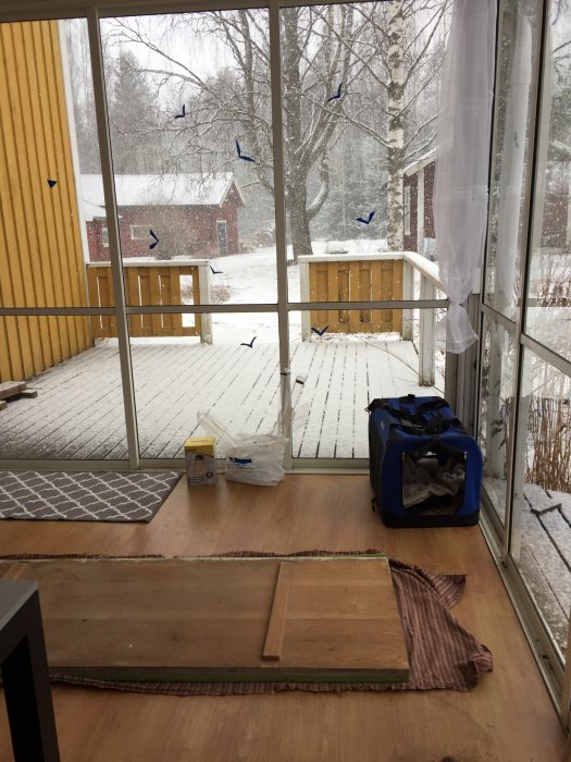 Uterum med en omklädd kökssoffa och utsikt över snöfall samt en trädgård med ett rött hus och nakna träd.