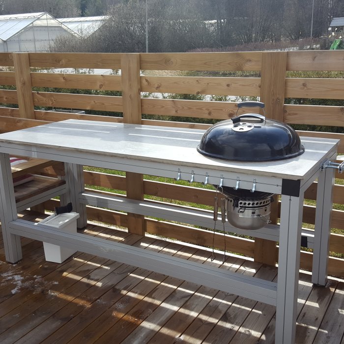Trägrillbord med en stenbänkskiva och en svart klotgrill på en trädäck terrass, med lite sot nära grillen.