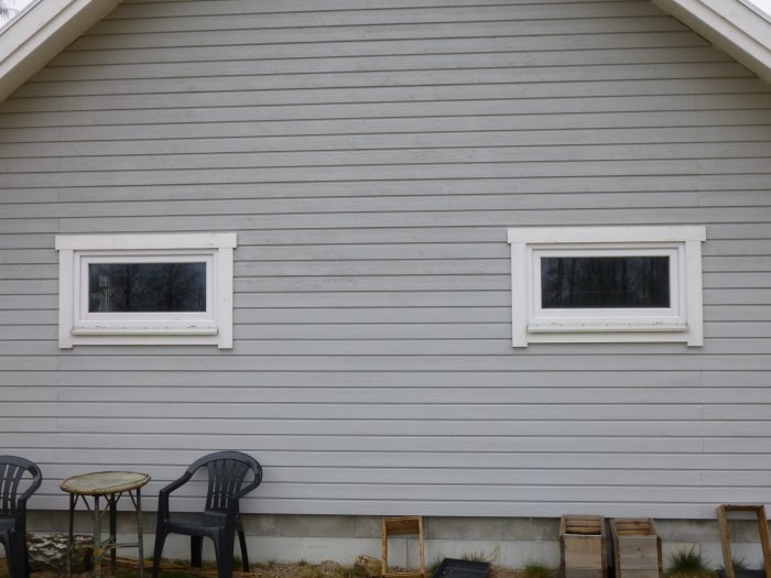 Två vita fönster på en husvägg med liggande träpanel och trädgårdsmöbler nedanför.