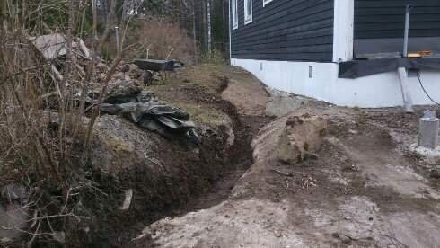Dräneringsarbete längs hus med berghäll, fiberduk och grus, grävmaskin i bakgrunden.