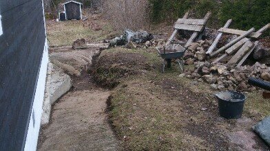 Grävd ränna längs med hus för dräneringsprojekt, med stenar, skottkärra och hink i trädgård.