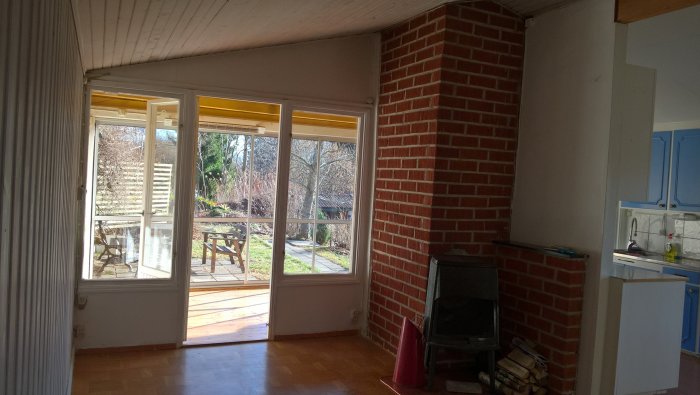 Inomhusvy av ett vardagsrum med stor glasdörr ut mot en trädgård, ett tegelskorsten med kamin, trägolv och del av kök.