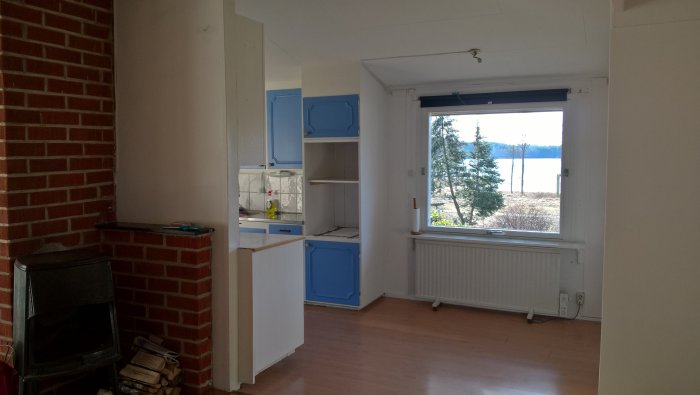 Interiör av ett vardagsrum med blick mot ett kök och ett fönster med sjöutsikt, kamin i förgrunden.