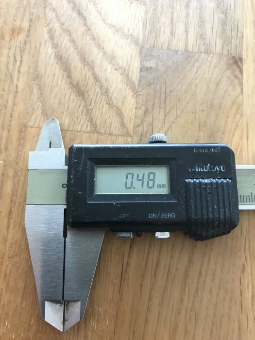 Digital skjutmått som visar en mätning på 0.48 mm mot en träbakgrund.