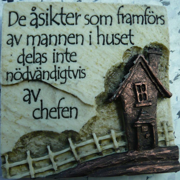 Dekorativ plakett med texten "De åsikter som framförs av mannen i huset delas inte nödvändigtvis av chefen" och en relief av ett hus.