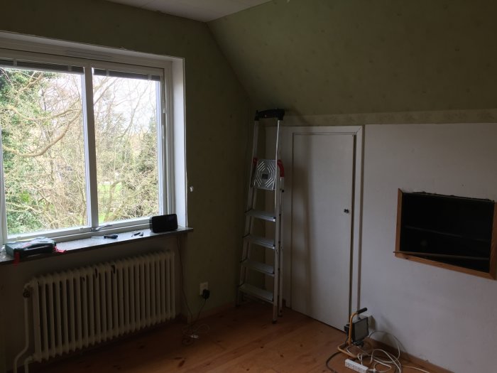 Oinredda rummet före renovering med tapetserade väggar, fönster, stege och eluttag.