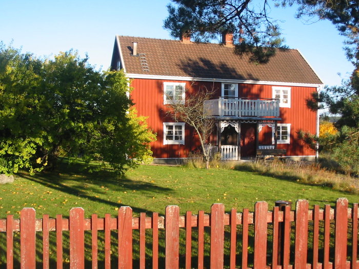 Rött trähus med vita knutar och balkong, omgivet av grönska och ett rött staket.