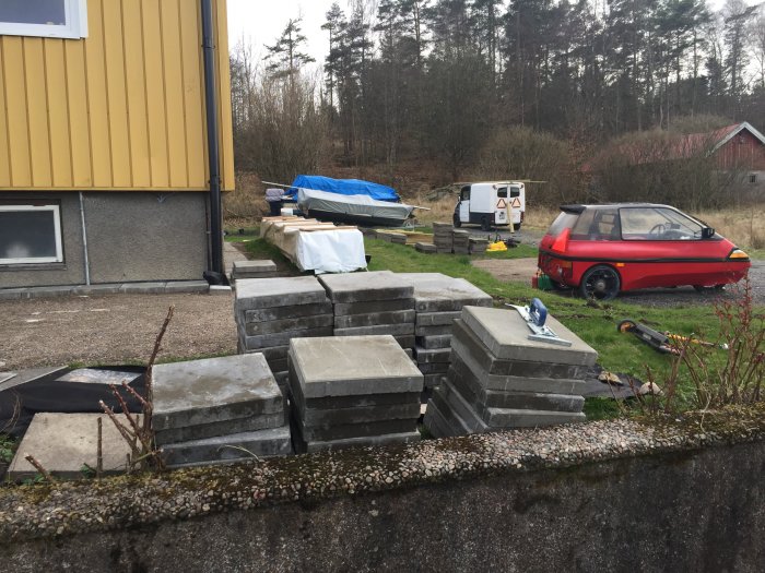 Staplade betongplattor på en trädgård vid sidan av ett hus, med byggmaterial och verktyg utspridda runtomkring.