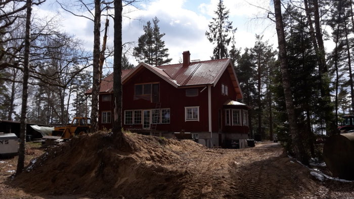 Rödfärgat hus med röd tegeltak i en skogsmiljö under renovering med byggmaterial och grävmaskin synliga.