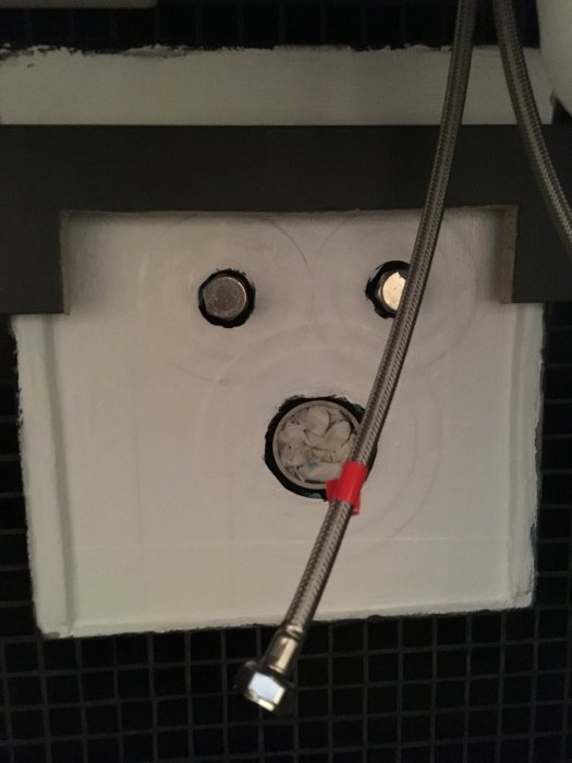 Två oisolerade kopparrör som sticker ut från en vit vägg med en svart kakelkant och en vattenledningsslang.