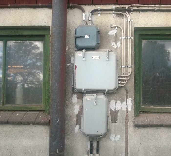 Äldre elutrustning monterad på en stenvägg vid en byggnad, inklusive stålrör och grå elektricitetskåpor.