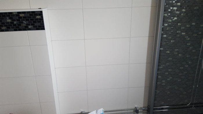 Vägg med stor vit matt kakelplatta vid en dusch och liten svart blank mosaik.