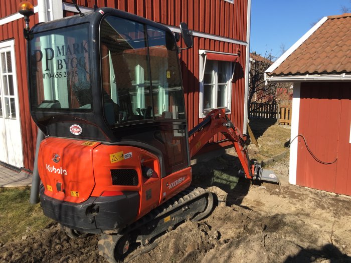 Minigrävare som arbetar på en trång byggplats mellan röda trähus.