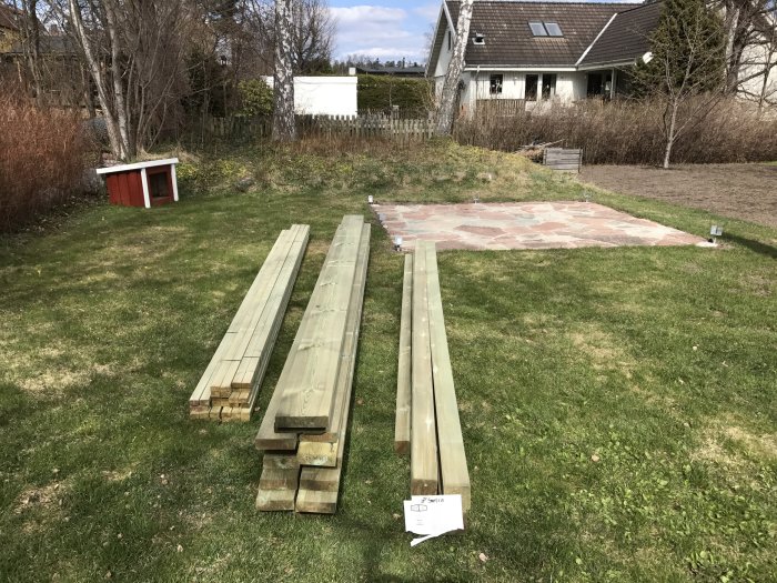 Träreglar inhandlade för byggprojekt ligger på en gräsmatta med ett hus och en trädgård i bakgrunden.