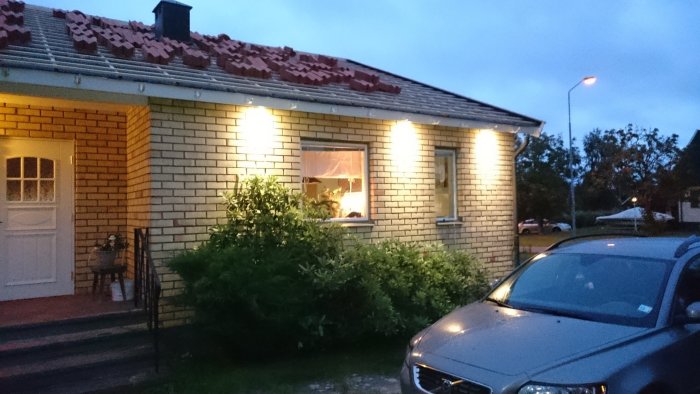 Ett hus med spotlights i takfoten som lyser upp fasaden på kvällen, med en bil parkerad framför.