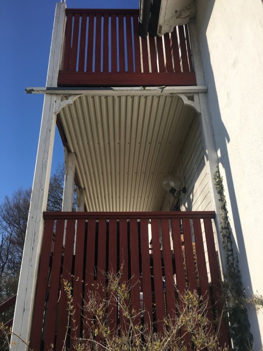 Liten sliten veranda på ett äldre hus med reveterad fasad och en balkong ovanpå.