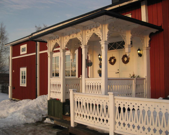 Tvåvåningshus med röda väggar och vit snickarglädje på veranda i vintersol.