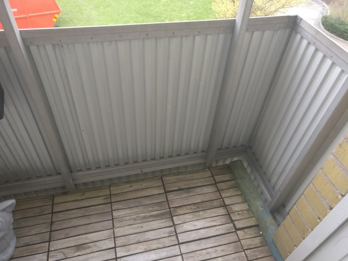 En delvis skyddad trädäck med lutande tak av korrugerad plåt och trästaket, vid sidan av en gul tegelbyggnad.