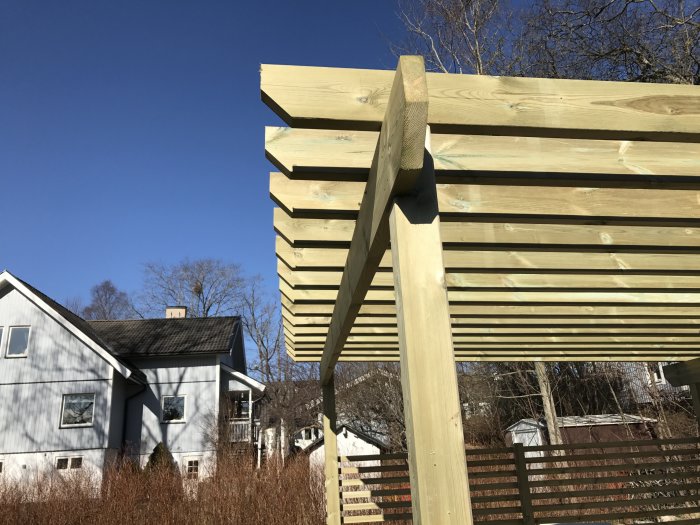 Del av en pergola i träkonstruktion med solida stolpar och horisontella takreglar mot en klarblå himmel.