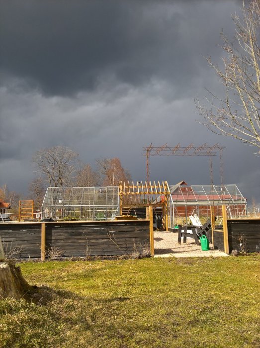 Trädgård med växthus under mörka moln, indikerande längtan till vårens grönska.