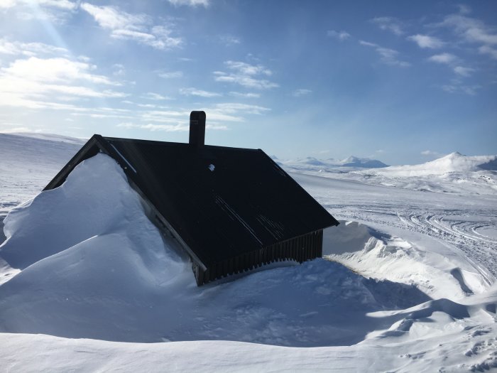 Fjällstuga delvis täckt av snö med spår från skotrar i det omgivande snötäckta landskapet.