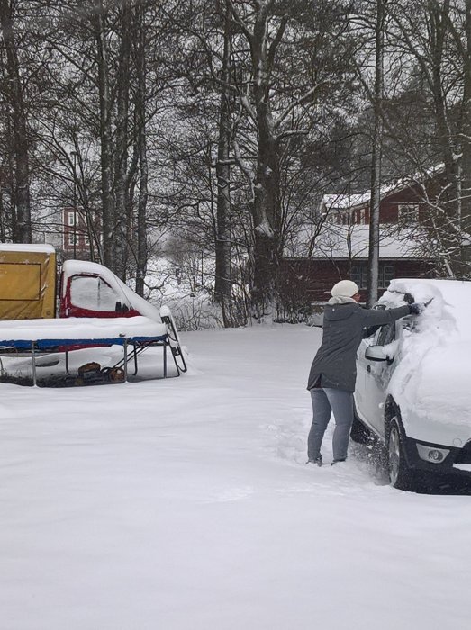 Person som borstar snö från bil framför vintertäckt hus och trädgård med tung snöfall.