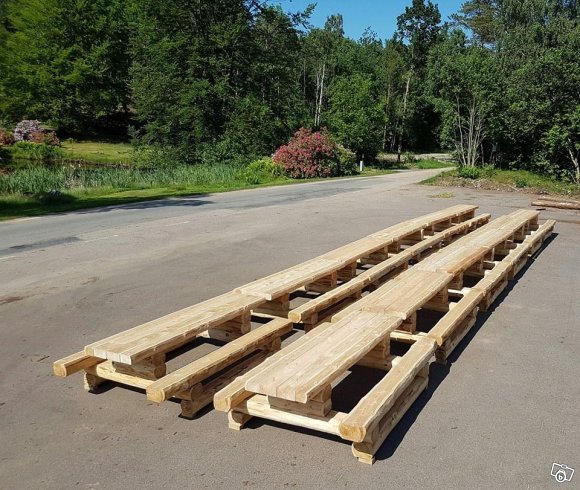 Staplade grovyxade timmerbord av tall på en asfalterad yta med grönska i bakgrunden.