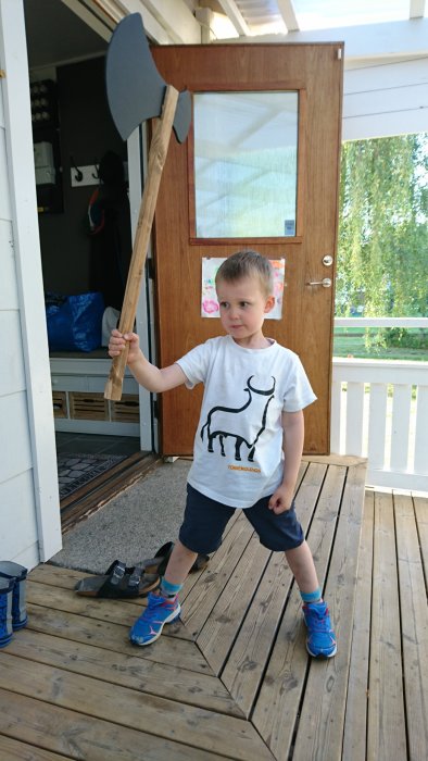 Pojke i vit t-shirt står på en träveranda med en handgjord träyxa, inspirerad av vikingatema.